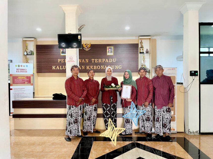 Group of six people in matching red traditional outfits stand in a government office lobby, one woman in green hijab holds a document while another holds a decorative basket and gift.