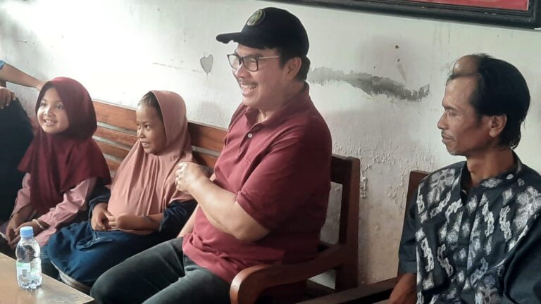 People sitting on a bench indoors, smiling and chatting; a man in a maroon polo and cap center, two women in hijabs on the left, and a man in a patterned shirt on the right. A water bottle sits on the table in front.