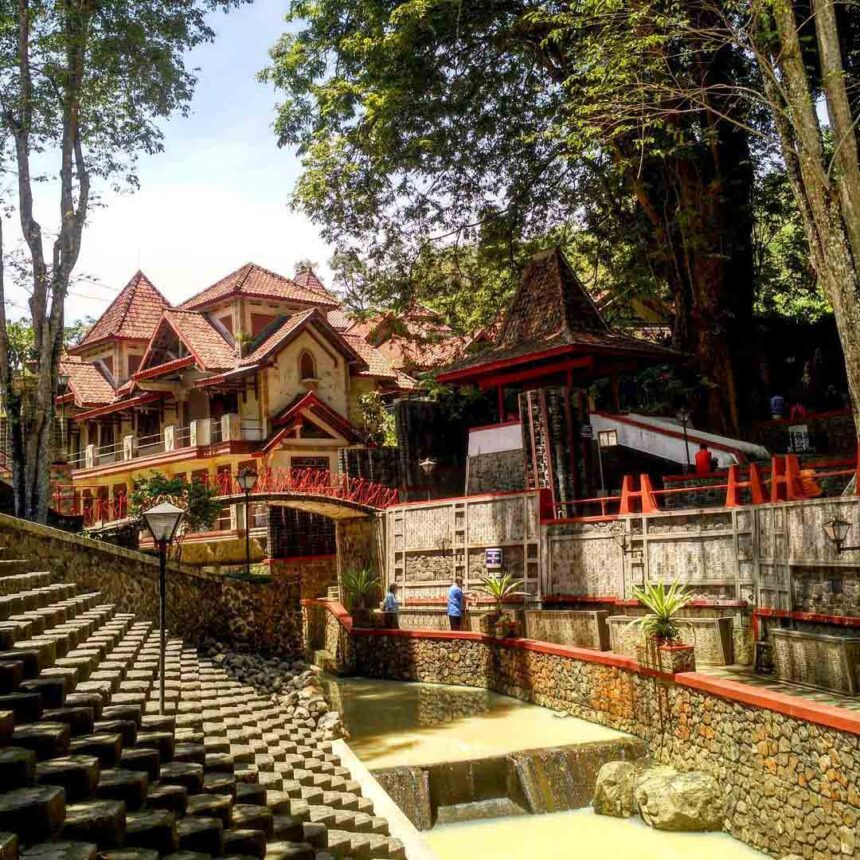 Cluster of traditional-style buildings with red-tiled roofs beside a stone stairway and a shallow canal in a shaded temple complex