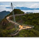 Suspension skywalk extending over a forested canyon with distant blue sea.