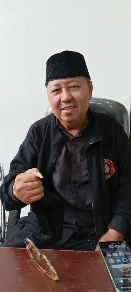 Smiling man in a black jacket and traditional cap sits at a desk, holding a small object in his right hand with glasses and a smartphone on the table nearby.