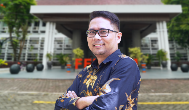 aSXA - Mabur.co Man wearing a blue batik shirt and glasses stands with arms crossed outside a modern building, smiling at the camera.