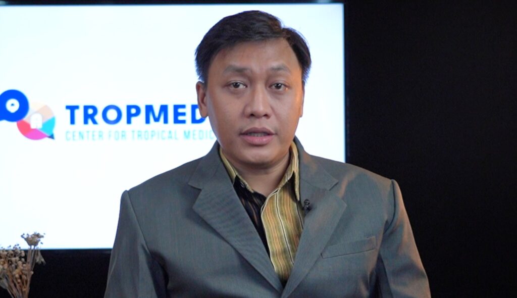 Man in a gray suit speaking in front of a screen that shows TROPMED Center for Tropical Medicine logo/text in blue.