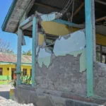 Exterior shot of a yellow-green building with heavy structural damage: crumbling walls, exposed concrete, and debris; two children stand nearby on the left under a sunny blue sky.