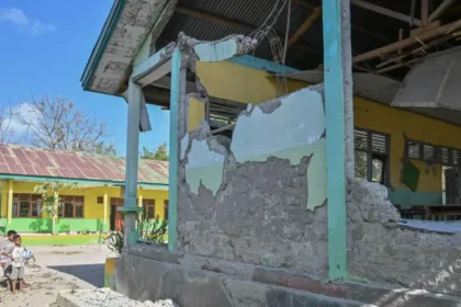 Exterior shot of a yellow-green building with heavy structural damage: crumbling walls, exposed concrete, and debris; two children stand nearby on the left under a sunny blue sky.