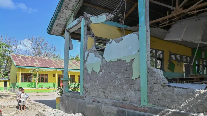 Exterior shot of a yellow-green building with heavy structural damage: crumbling walls, exposed concrete, and debris; two children stand nearby on the left under a sunny blue sky.