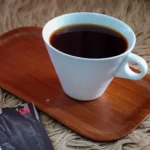 White coffee cup filled with dark coffee sits on a wooden tray on a shaggy beige rug, with a magazine partially visible in the corner.