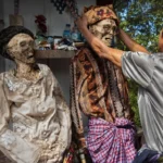 A man adjusts the head of a life-size skeletal figure wearing a patterned headwrap, with another dressed skeleton nearby in an outdoor, shrine-like setting.