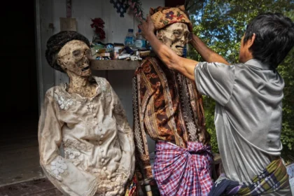 A man adjusts the head of a life-size skeletal figure wearing a patterned headwrap, with another dressed skeleton nearby in an outdoor, shrine-like setting.
