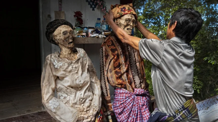 aXax - Mabur.co A man adjusts the head of a life-size skeletal figure wearing a patterned headwrap, with another dressed skeleton nearby in an outdoor, shrine-like setting.