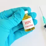 Gloved hand holding a small vial labeled HIV vaccine beside a syringe on a white surface, ready for administration.