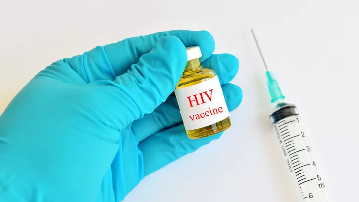 Gloved hand holding a small vial labeled HIV vaccine beside a syringe on a white surface, ready for administration.