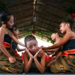Group of young girls in traditional batik outfits perform a coordinated dance under a thatched roof, with one girl seated center posing with hands to her cheeks.