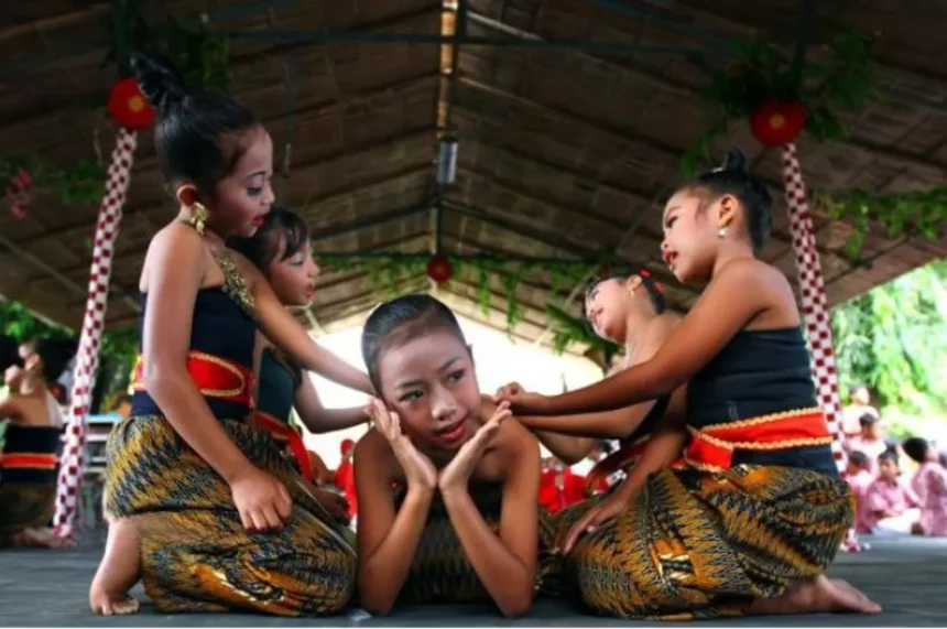 Group of young girls in traditional batik outfits perform a coordinated dance under a thatched roof, with one girl seated center posing with hands to her cheeks.