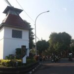 White guardhouse with red-roofed tower at a street intersection; motorcycles and pedestrians nearby under a curved streetlight.
