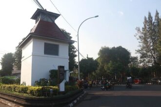 White guardhouse with red-roofed tower at a street intersection; motorcycles and pedestrians nearby under a curved streetlight.