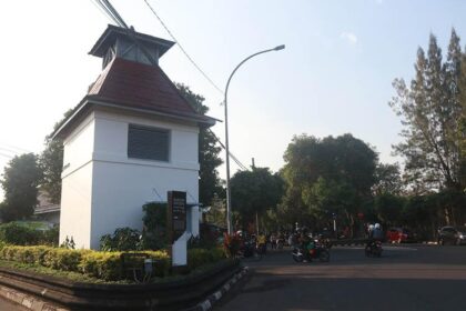 White guardhouse with red-roofed tower at a street intersection; motorcycles and pedestrians nearby under a curved streetlight.