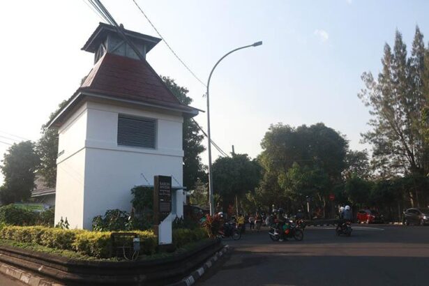 White guardhouse with red-roofed tower at a street intersection; motorcycles and pedestrians nearby under a curved streetlight.
