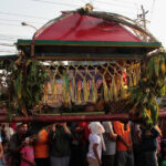People in colorful clothing carry a decorated palanquin wrapped with leaves and a red canopy during a festival.