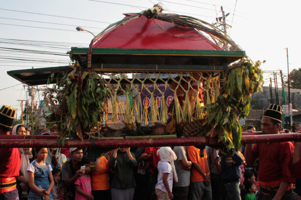 People in colorful clothing carry a decorated palanquin wrapped with leaves and a red canopy during a festival.