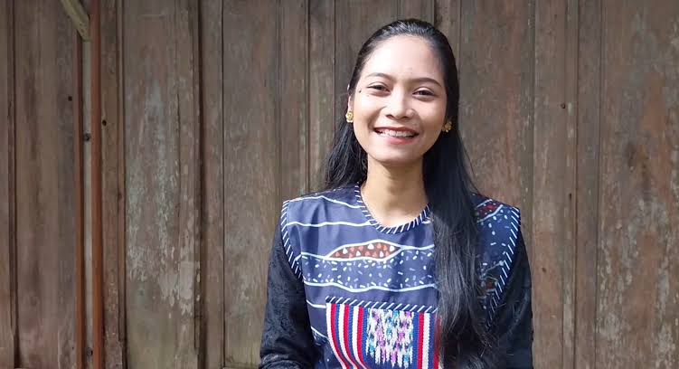 aZza - Mabur.co Smiling woman with long black hair wearing a blue patterned traditional top, standing in front of a wooden fence.