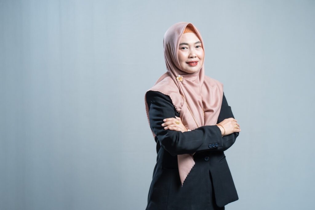 aZza - Mabur.co Portrait of a woman in a pink hijab and black blazer, arms crossed, smiling softly against a neutral background.