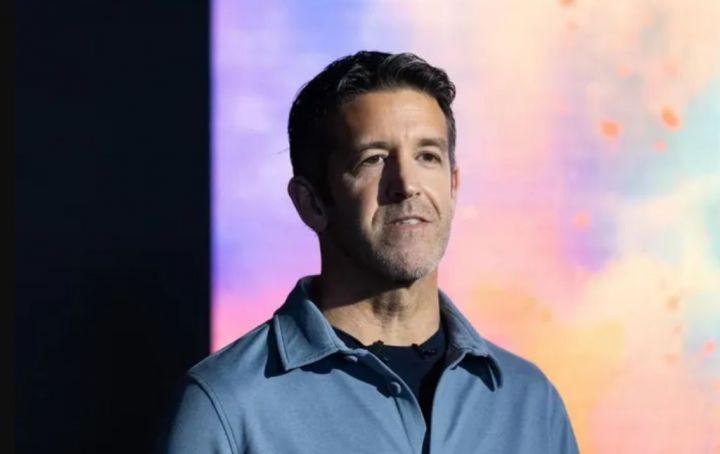 Male speaker in a blue collared shirt on stage with a colorful abstract background, delivering a talk