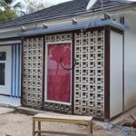 Small white, single-story house with a decorative concrete lattice screen over the red front door, in a dirt yard with a rooster nearby.