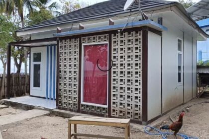 Small white, single-story house with a decorative concrete lattice screen over the red front door, in a dirt yard with a rooster nearby.