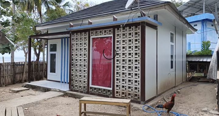 Small white, single-story house with a decorative concrete lattice screen over the red front door, in a dirt yard with a rooster nearby.