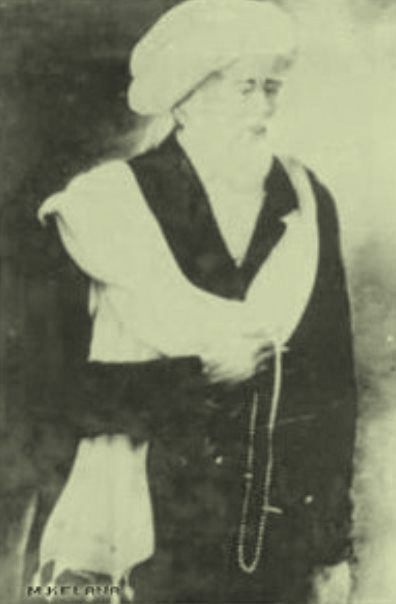 Historical black-and-white portrait of a person wearing a turban and dark robe with a light shawl, holding a bead necklace.