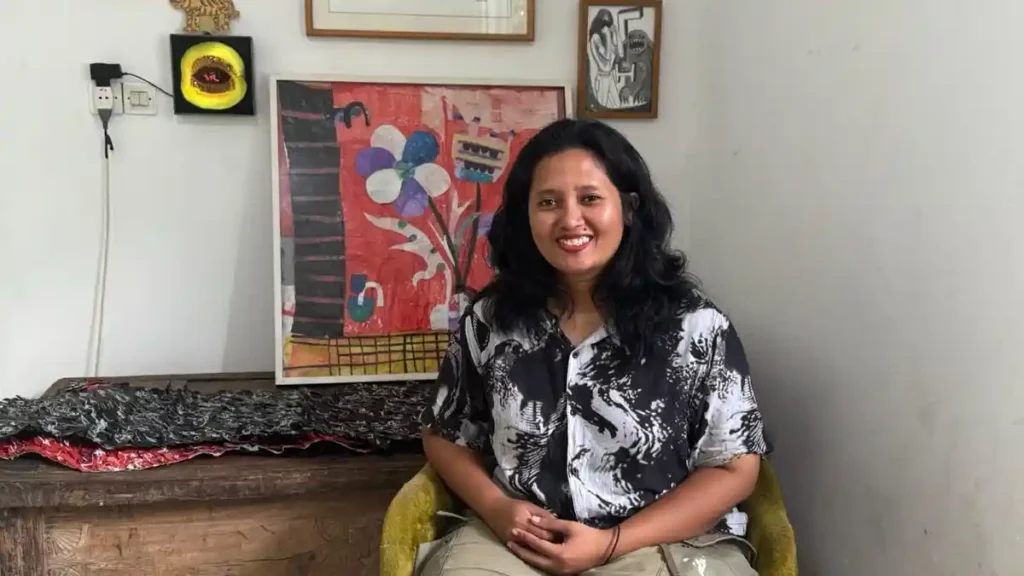 acTic - Mabur.co Smiling woman with dark hair sits in a yellow chair, wearing a black-and-white patterned shirt, with colorful artwork behind her.