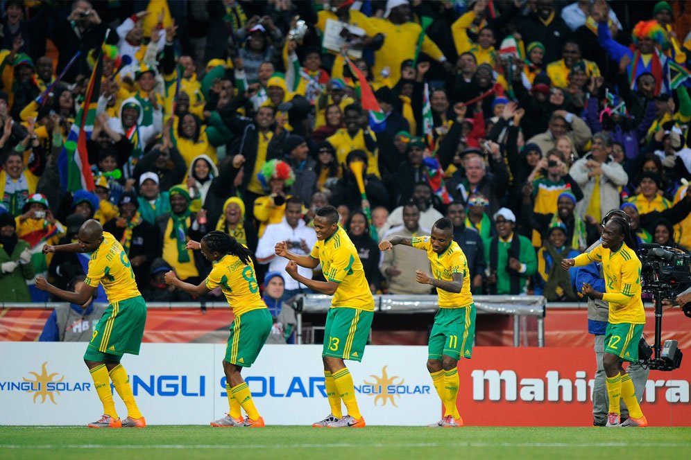 Soccer players in yellow and green celebrate a goal on the field as a cheering crowd watches the scene in the stands.