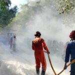 Firefighters in orange and blue protective gear spraying water through a hose in a smoke-filled street during a fire event.