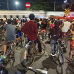 A large group of cyclists standing with their bikes on a nighttime city street as a train whizzes by in the background, with crossing signs visible nearby.