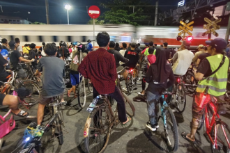 A large group of cyclists standing with their bikes on a nighttime city street as a train whizzes by in the background, with crossing signs visible nearby.