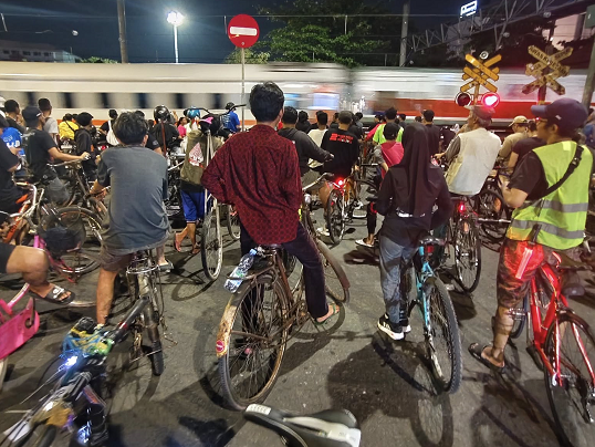 A large group of cyclists standing with their bikes on a nighttime city street as a train whizzes by in the background, with crossing signs visible nearby.