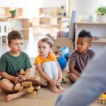 Four preschoolers sit on a beige carpet in a bright classroom, listening to a teacher offscreen.