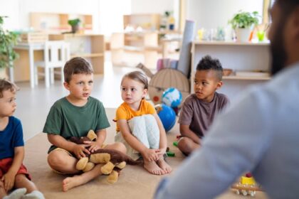 Four preschoolers sit on a beige carpet in a bright classroom, listening to a teacher offscreen.