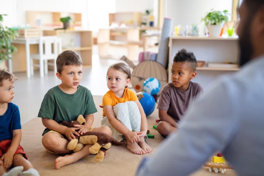 Four preschoolers sit on a beige carpet in a bright classroom, listening to a teacher offscreen.