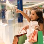 Mother and young girl shopping in a mall, the child pointing at a store window display while carrying gift bags.