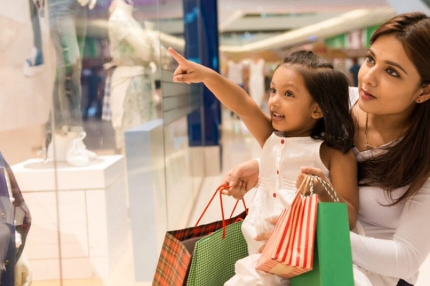 Mother and young girl shopping in a mall, the child pointing at a store window display while carrying gift bags.