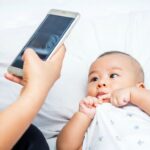 Baby lying on a bed looks at a smartphone held by an adult nearby.