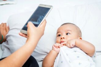Baby lying on a bed looks at a smartphone held by an adult nearby.