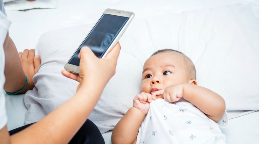 Baby lying on a bed looks at a smartphone held by an adult nearby.