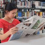 Woman in a red shirt reads a newspaper at a library table, with tall bookshelves behind her.
