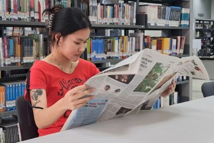 Woman in a red shirt reads a newspaper at a library table, with tall bookshelves behind her.