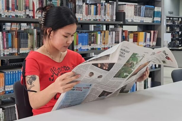 Woman in a red shirt reads a newspaper at a library table, with tall bookshelves behind her.