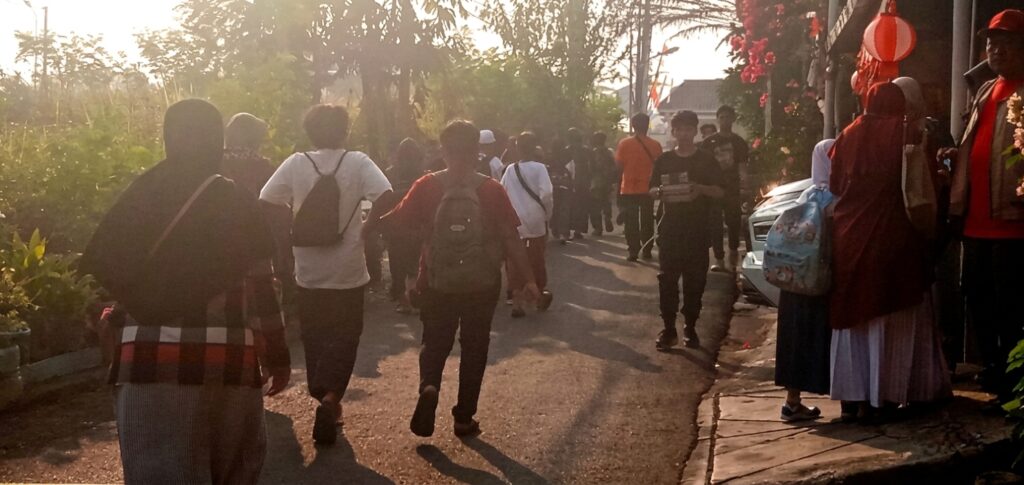 anak2 jalan ke bus FIXX - Mabur.co People walking down a sunlit street in a casual procession, several with backpacks and hoodies. Left side is vegetation; right side has onlookers in festive dress near a car.