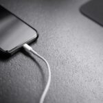 Close-up of a smartphone on a textured surface with a white charging cable connected to its bottom edge.